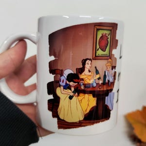 Könnte beinhalten: Weißer Keramikbecher mit einer humorvollen Illustration von Disney-Prinzessinnen. Das Bild zeigt Schneewittchen, Belle und Aschenputtel an einem Tisch mit Weinflaschen. Ein gerahmtes Bild eines Herzens befindet sich an der Wand. Der Becher hat einen gebogenen Griff.