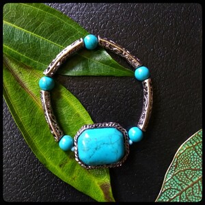 May include: A turquoise gemstone bracelet with a large, rectangular turquoise stone in the center. The bracelet is made of silver-toned metal with a decorative design. The bracelet is on a green leaf background.