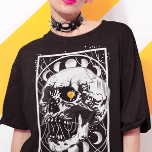 K&ouml;nnte beinhalten: Schwarzes T-Shirt mit einem grafischen Druck eines wei&szlig;en Sch&auml;dels mit einem gelben Auge, umgeben von Monden und Sternen. Der Sch&auml;del befindet sich in einem schwarzen quadratischen Rahmen.