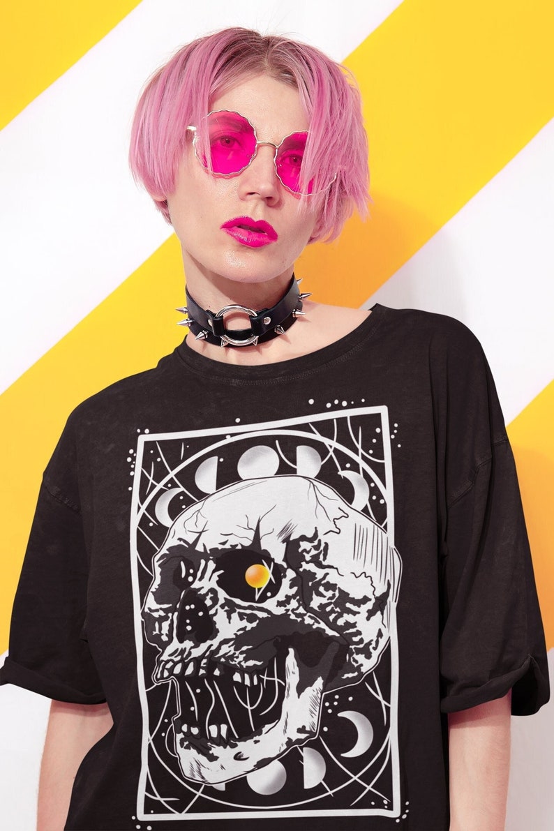 K&ouml;nnte beinhalten: Schwarzes T-Shirt mit einem grafischen Druck eines wei&szlig;en Sch&auml;dels mit einem gelben Auge, umgeben von Monden und Sternen. Der Sch&auml;del befindet sich in einem schwarzen quadratischen Rahmen.