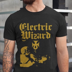 Puede incluir: Una camiseta negra con un estampado gráfico dorado del texto "Electric Wizard" en una fuente gótica. El gráfico también incluye una cruz estilizada y una lágrima.