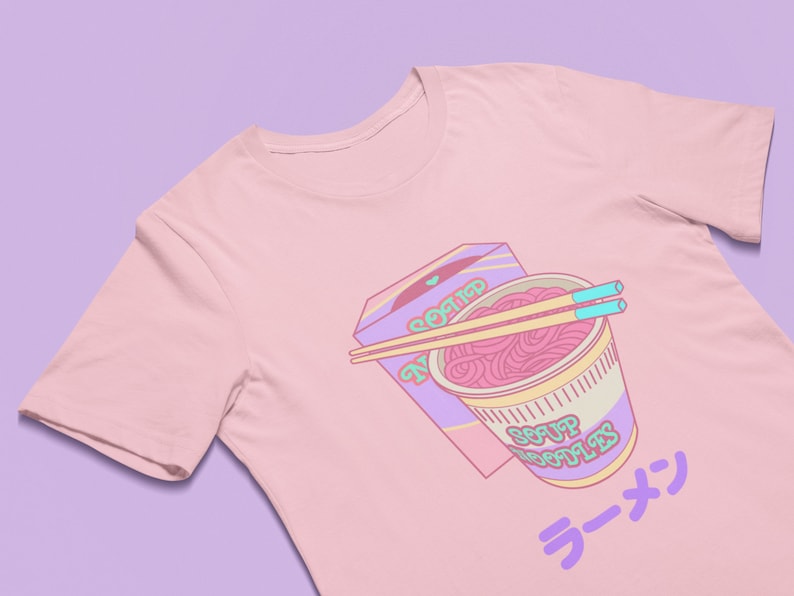 May include: A pink t-shirt with a graphic of a bowl of ramen noodles, chopsticks, and a box that says "Noodle" on it. The text "Soup Noodles" is on the bowl and "ラーメン" is on the bottom of the shirt.