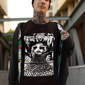 Puede incluir: Una camiseta marr&oacute;n con un estampado gr&aacute;fico en blanco y negro de una cara con el texto japon&eacute;s "森田のかげ" en la parte delantera. La persona que lleva la camiseta lleva una boina negra, un aro negro en la nariz y tatuajes en el cuello.