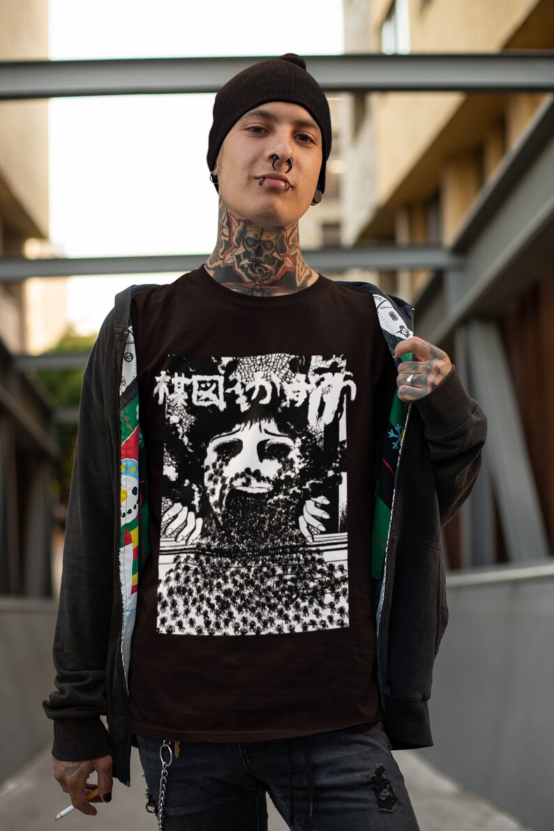 Puede incluir: Una camiseta marr&oacute;n con un estampado gr&aacute;fico en blanco y negro de una cara con el texto japon&eacute;s "森田のかげ" en la parte delantera. La persona que lleva la camiseta lleva una boina negra, un aro negro en la nariz y tatuajes en el cuello.