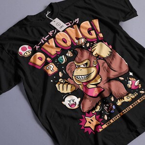 Puede incluir: Camiseta negra con un diseño de Donkey Kong y el texto "D. Kong!" en naranja y rosa. El diseño incluye texto japonés e imágenes de la película Super Mario Bros.