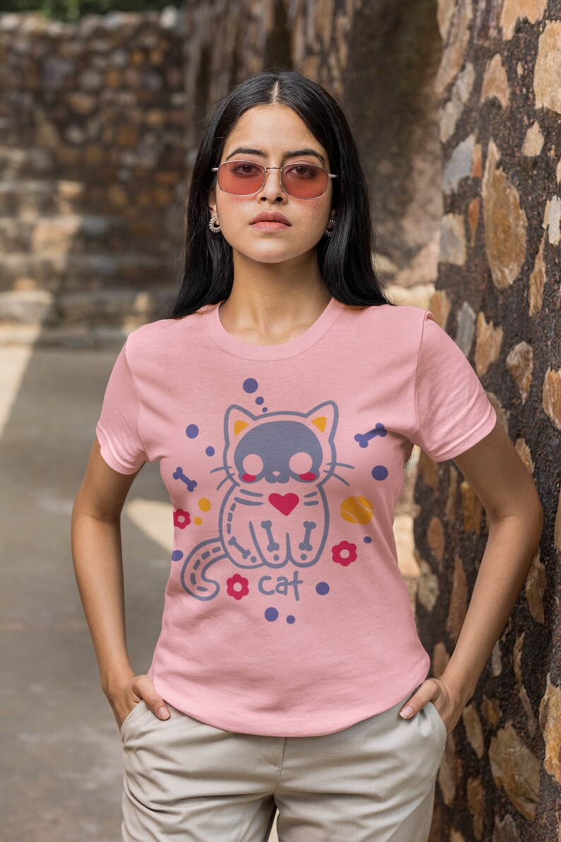 Peut inclure: Un t-shirt rose avec un motif de chat de dessin anim&eacute;. Le chat porte un c&oelig;ur et a des os pour les membres. Le texte "I cat" est imprim&eacute; sous le chat.