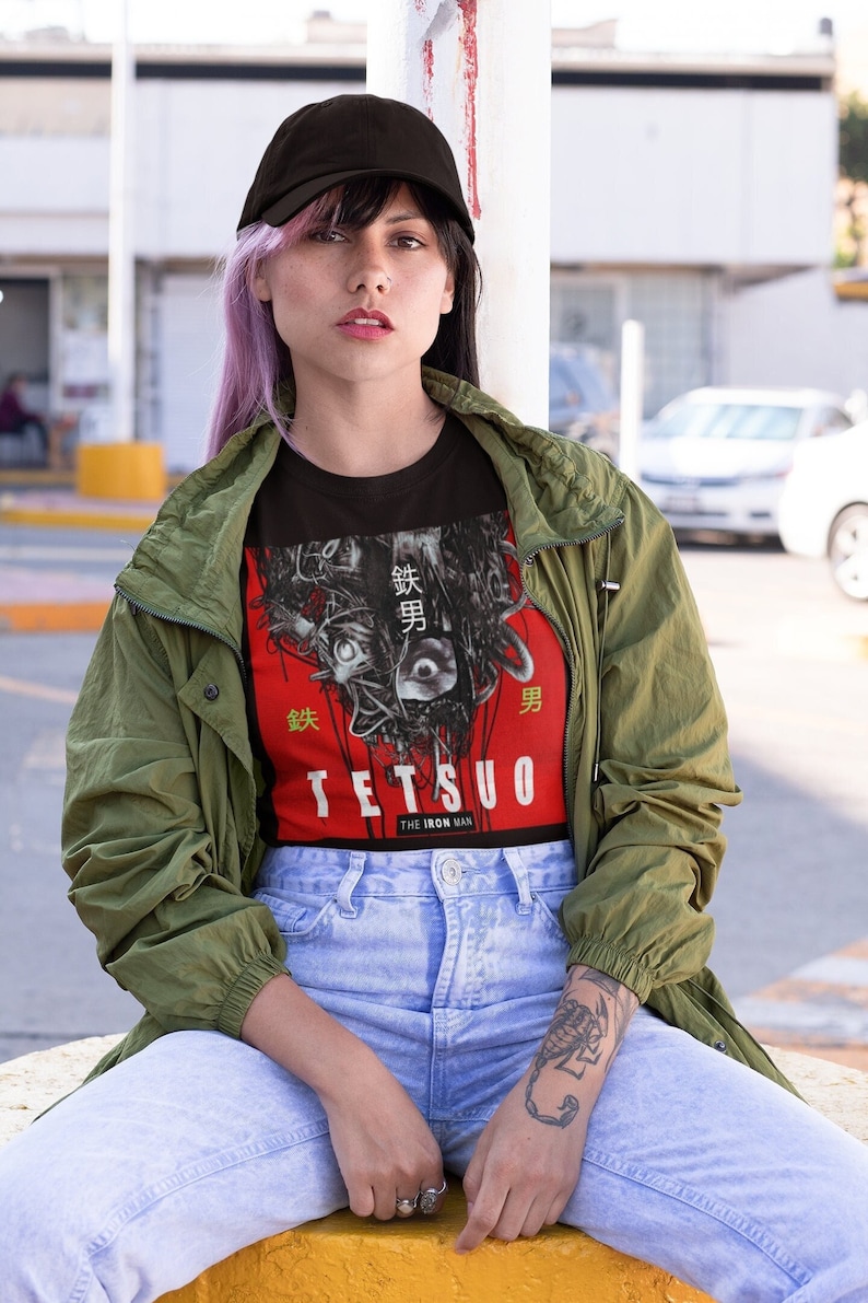 Puede incluir: Una persona lleva una camiseta negra con un estampado gr&aacute;fico de una cara de cyborg y el texto "Tetsuo The Iron Man" en japon&eacute;s. La persona tambi&eacute;n lleva una gorra de b&eacute;isbol negra, una chaqueta verde y unos vaqueros azules.