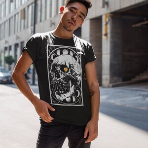 K&ouml;nnte beinhalten: Ein schwarzes T-Shirt mit einem wei&szlig;en Grafikdruck eines Sch&auml;dels mit einem Auge in der Mitte. Der Sch&auml;del ist von einem Kreis aus Monden und Sternen umgeben. Der Text "Cosmic" befindet sich &uuml;ber dem Sch&auml;del.