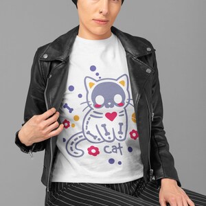 Peut inclure: T-shirt blanc avec un motif de chat violet avec un design de squelette et le mot "cat" imprim&eacute; en dessous. Le chat porte un c&oelig;ur rouge. Le chat est entour&eacute; d'os jaunes, de fleurs rouges et de points blancs.