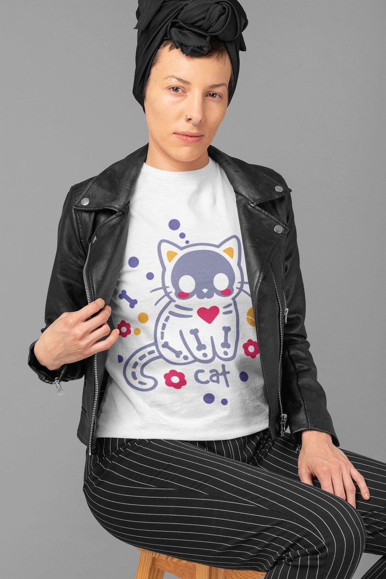 Peut inclure: T-shirt blanc avec un motif de chat violet avec un design de squelette et le mot "cat" imprim&eacute; en dessous. Le chat porte un c&oelig;ur rouge. Le chat est entour&eacute; d'os jaunes, de fleurs rouges et de points blancs.