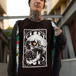 K&ouml;nnte beinhalten: Ein schwarzes T-Shirt mit einem wei&szlig;en Grafikdruck eines Sch&auml;dels mit einem gelben Auge, umgeben von Monden und Sternen. Der Sch&auml;del befindet sich in einem wei&szlig;en Rahmen.