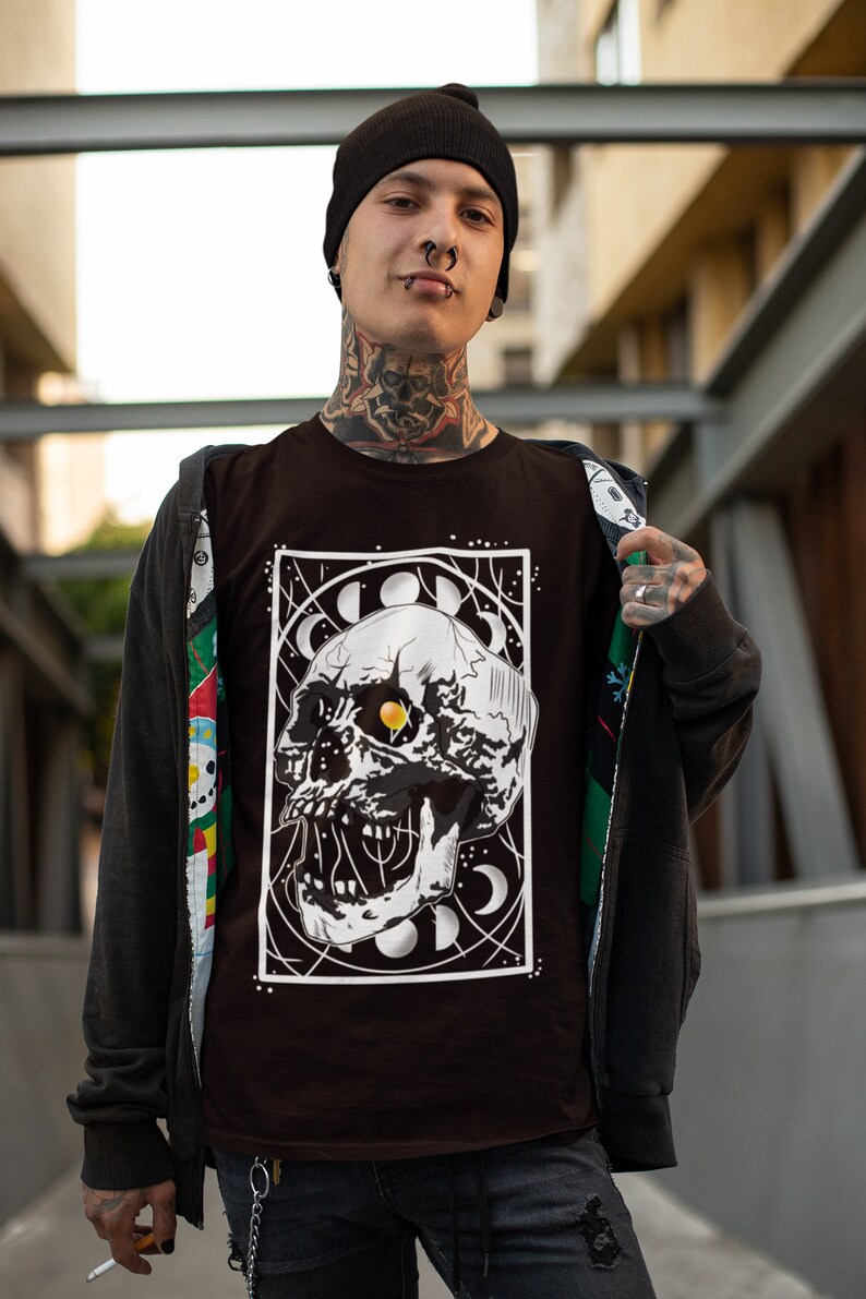 K&ouml;nnte beinhalten: Ein schwarzes T-Shirt mit einem wei&szlig;en Grafikdruck eines Sch&auml;dels mit einem gelben Auge, umgeben von Monden und Sternen. Der Sch&auml;del befindet sich in einem wei&szlig;en Rahmen.