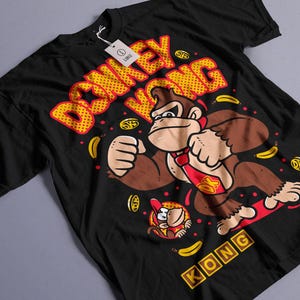 Puede incluir: Camiseta negra con un diseño de Donkey Kong, mostrando el texto "DONKEY KONG" en amarillo y rojo. El diseño incluye imágenes de plátanos y monedas. También aparece un personaje de Donkey Kong más pequeño.