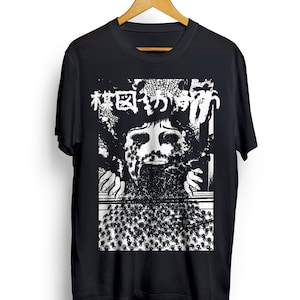 Puede incluir: Camiseta negra con un estampado gr&aacute;fico blanco de una cara rodeada de ara&ntilde;as. El texto "Bosque de ara&ntilde;as" est&aacute; impreso en caracteres japoneses sobre la cara.
