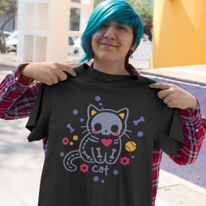 Peut inclure: T-shirt noir avec une image de dessin anim&eacute; d'un chat squelette tenant un c&oelig;ur rouge. Le chat est entour&eacute; de fleurs violettes, d'os et d'une balle de tennis jaune. Le texte "cat" est imprim&eacute; sous le chat.