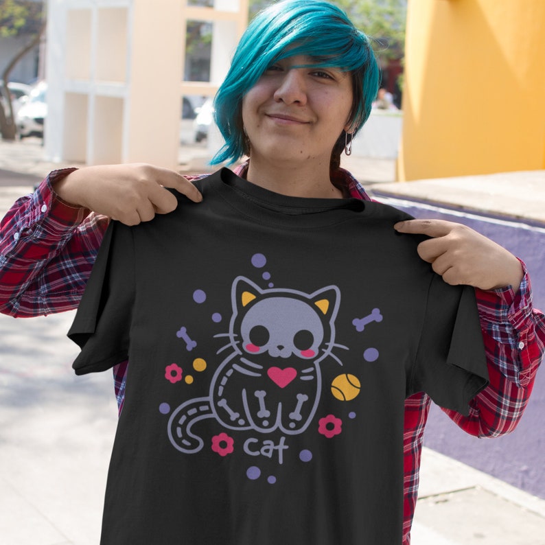 Peut inclure: T-shirt noir avec une image de dessin anim&eacute; d'un chat squelette tenant un c&oelig;ur rouge. Le chat est entour&eacute; de fleurs violettes, d'os et d'une balle de tennis jaune. Le texte "cat" est imprim&eacute; sous le chat.