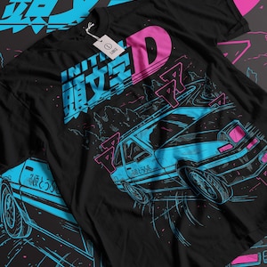 Puede incluir: Camiseta negra con un diseño gráfico azul y rosa. El diseño presenta un coche de carreras en una carretera de montaña, con texto japonés y la letra "D". Una etiqueta está adherida a la camiseta.