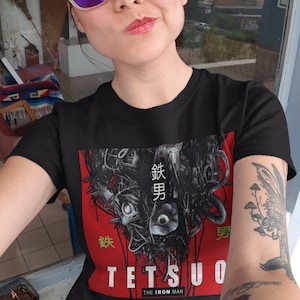 Puede incluir: Una persona lleva una camiseta negra con un estampado gr&aacute;fico de un robot con texto japon&eacute;s que dice "鉄男" (Tetsuo) y "The Iron Man".