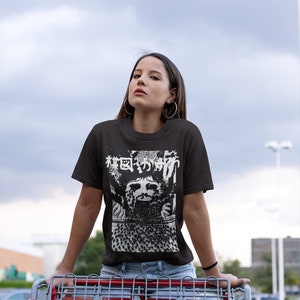 Puede incluir: Una mujer con una camiseta negra con un estampado gr&aacute;fico blanco de una cara y texto japon&eacute;s. El texto dice "Bosque de los Muertos". Ella est&aacute; de pie frente a un carrito de compras.
