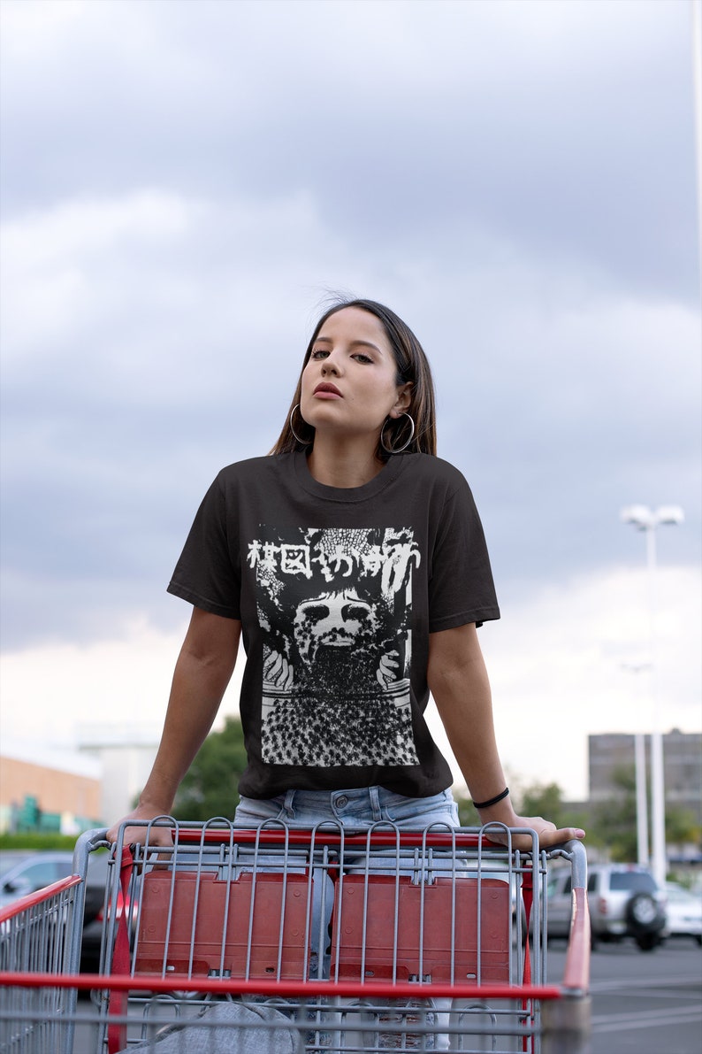 Puede incluir: Una mujer con una camiseta negra con un estampado gr&aacute;fico blanco de una cara y texto japon&eacute;s. El texto dice "Bosque de los Muertos". Ella est&aacute; de pie frente a un carrito de compras.