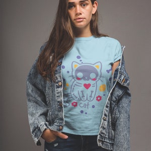 Peut inclure: Une femme portant un t-shirt bleu clair avec un motif de chat de dessin anim&eacute; avec un cr&acirc;ne et des os crois&eacute;s. Le chat tient un c&oelig;ur et a des points de suture sur son corps. Le texte "Cat" est sous le chat. La femme porte &eacute;galement une veste en jean et un jean bleu.