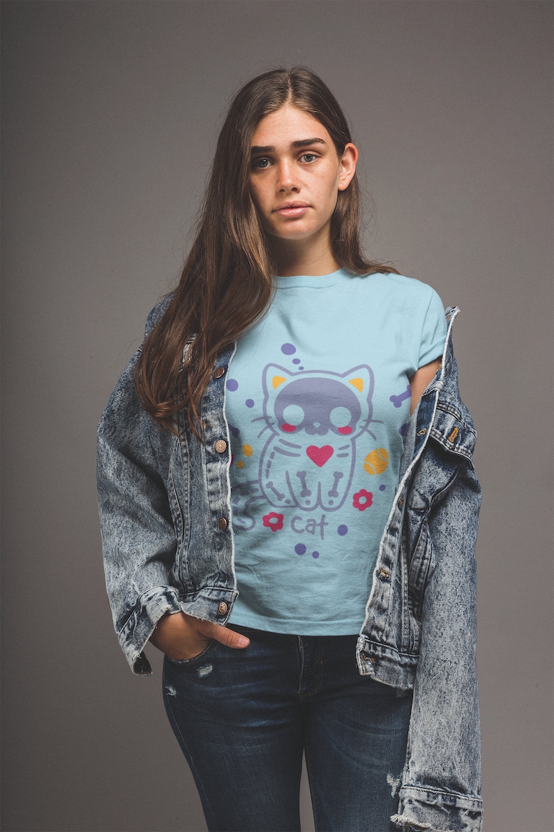 Peut inclure: Une femme portant un t-shirt bleu clair avec un motif de chat de dessin anim&eacute; avec un cr&acirc;ne et des os crois&eacute;s. Le chat tient un c&oelig;ur et a des points de suture sur son corps. Le texte "Cat" est sous le chat. La femme porte &eacute;galement une veste en jean et un jean bleu.