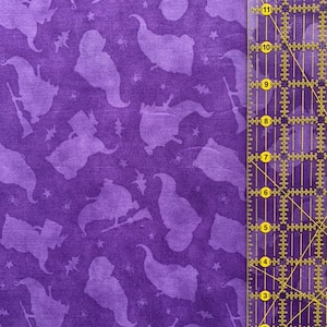 May include: Purple fabric with a repeating pattern of witches flying on broomsticks. The fabric is textured and has a subtle sheen. The witches are silhouetted and are flying against a background of stars and spiders.