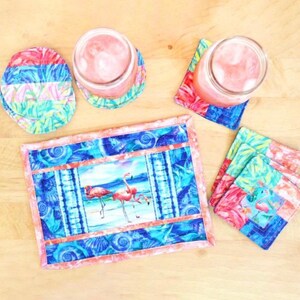 May include: A set of four pink flamingo themed coasters with a matching placemat. The coasters and placemat feature a blue and pink colour scheme with a seashell pattern. The placemat has a picture of two pink flamingos standing in the water.