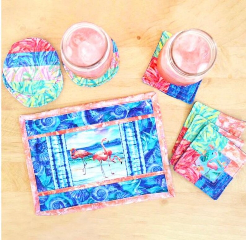 May include: A set of four pink flamingo themed coasters with a matching placemat. The coasters and placemat feature a blue and pink colour scheme with a seashell pattern. The placemat has a picture of two pink flamingos standing in the water.