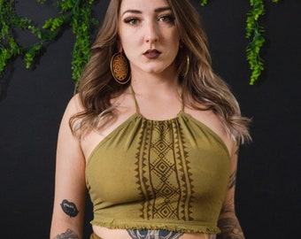 May include: A woman wearing a green halter top with a brown geometric pattern and a brown wrap skirt with a similar pattern. She has long brown hair and is wearing large gold hoop earrings.