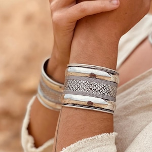 Op de afbeelding: Twee zilveren armbanden om de pols van een persoon. De armband bovenaan heeft een getextureerd ontwerp en de armband onderaan is effen.
