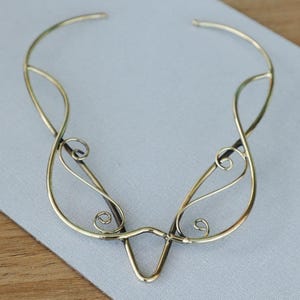 Può includere: Una delicata collana in metallo dorato con un design aperto. La collana presenta linee curve, volute e una forma a V centrale. Il pezzo è esposto su una superficie grigio chiaro, che mette in risalto i suoi dettagli intricati.