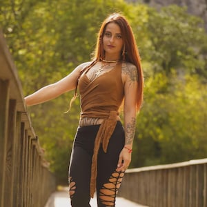 Könnte beinhalten: Ein braunes ärmelloses Wickeloberteil mit Bindedetail an der Taille, kombiniert mit einer schwarzen Leggings, die aufwendige seitliche Ausschnitte mit Kreuzschnürung aufweist. Das Outfit wird durch schwarze Plateausandalen abgerundet.