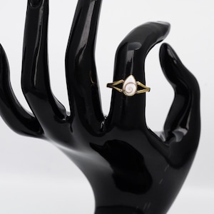 Puede incluir: Un anillo dorado con un diseño de concha blanca y beige en forma de lágrima. El anillo se exhibe en un soporte de joyería negro con forma de mano sobre un fondo blanco. La banda del anillo tiene un diseño dividido.
