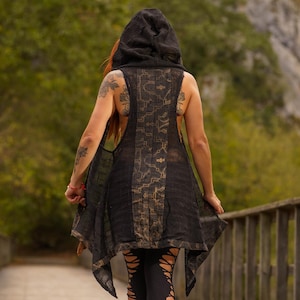 May include: A person wearing a black hooded vest with a patterned design, black leggings with cutouts, and sandals, walking on a wooden bridge. The vest has a unique, open-back design.
