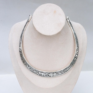 May include: A silver-toned metal collar necklace featuring an open-ended design. The surface of the necklace is decorated with intricate embossed patterns of small flowers and leaves, with a textured border running along the inner and outer edges.
