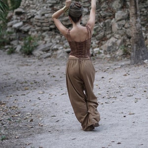 May include: A woman wearing a brown top with a laced back and brown pants, standing in front of a stone wall with her arms raised.