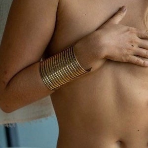 May include: A gold-toned bangle bracelet made of multiple stacked rings. The bracelet is worn on the wrist of a person.