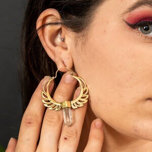May include: Gold hoop earrings with a clear quartz crystal pendant. The earrings have a delicate, intricate design with a wing-like pattern.