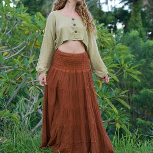 May include: A woman wearing a two-piece outfit. The top is a light green, long-sleeved crop top with three buttons. The bottom is a long, tiered, rust-colored skirt. The outfit is worn outdoors in a tropical setting.