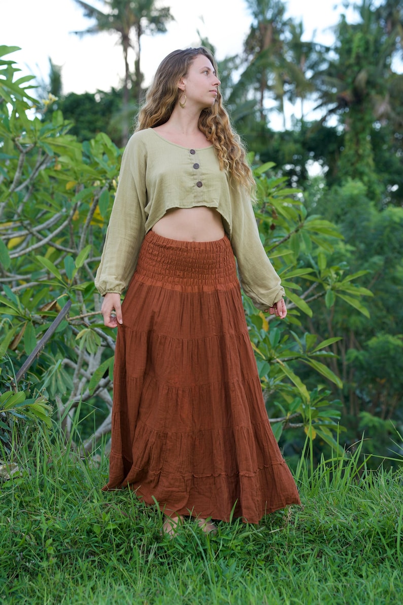 May include: A woman wearing a two-piece outfit. The top is a light green, long-sleeved crop top with three buttons. The bottom is a long, tiered, rust-colored skirt. The outfit is worn outdoors in a tropical setting.