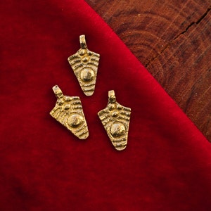 May include: Three gold-colored pendants with a textured, arrow-like design. Each pendant features a central circular element and smaller, raised details. The pendants rest on a deep red fabric, with a wooden surface in the background.