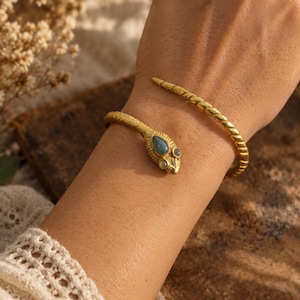 May include: A gold-toned metal cuff bracelet shaped like a coiled snake with a textured, scale-like surface. The snake head features a teardrop-shaped blue stone and a smaller round stone, while the tail end tapers into a smooth, twisted design.