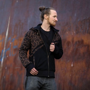 May include: A black zip-up hoodie with a brown patterned design. The hoodie has a hood and a zipper closure. The person is wearing the hoodie and has their hair pulled back in a bun.