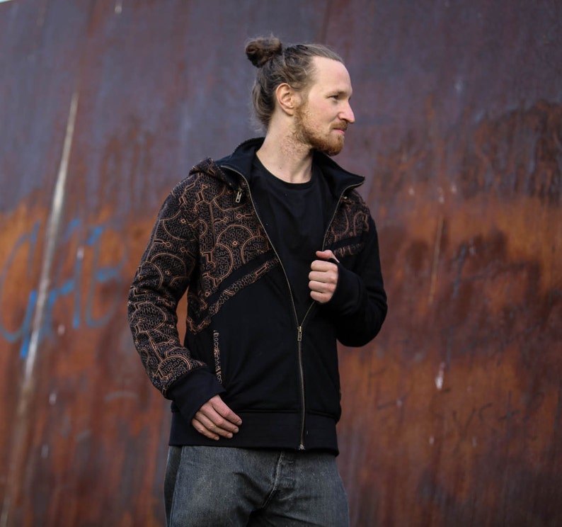 May include: A black zip-up hoodie with a brown patterned design. The hoodie has a hood and a zipper closure. The person is wearing the hoodie and has their hair pulled back in a bun.
