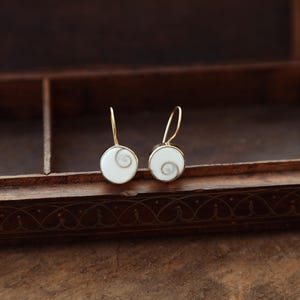 May include: A pair of gold-coloured drop earrings. Each earring has a white, circular pendant with a spiral design in the centre. The earrings are displayed on a wooden surface, highlighting their elegant design.