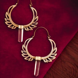 May include: A pair of gold hoop earrings with a clear crystal pendant hanging from each hoop. The earrings have a delicate, intricate design with a wing-like pattern.