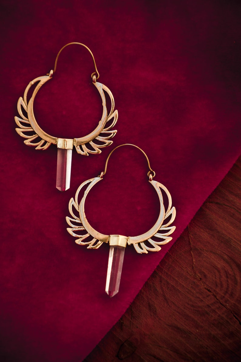 May include: A pair of gold hoop earrings with a clear crystal pendant hanging from each hoop. The earrings have a delicate, intricate design with a wing-like pattern.