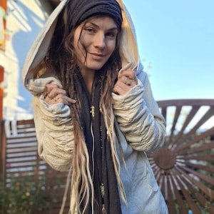 Könnte beinhalten: Hellbeige Kapuzenjacke mit gerafften Ärmeln. Die Jacke wird mit einem schwarzen Stirnband und Schal getragen. Die Person trägt Ringe. Der Hintergrund umfasst einen Holzzaun und einen blauen Himmel.