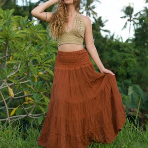 May include: A woman wearing a two-piece outfit outdoors. The top is a light green halter top with a geometric design. The skirt is a long, tiered, rust-colored skirt. The background features lush green foliage and palm trees.